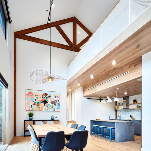 Main Ridge Barn dining high celing with timber trusses across dining table to kitchen with feature blue - building photographer example / concept